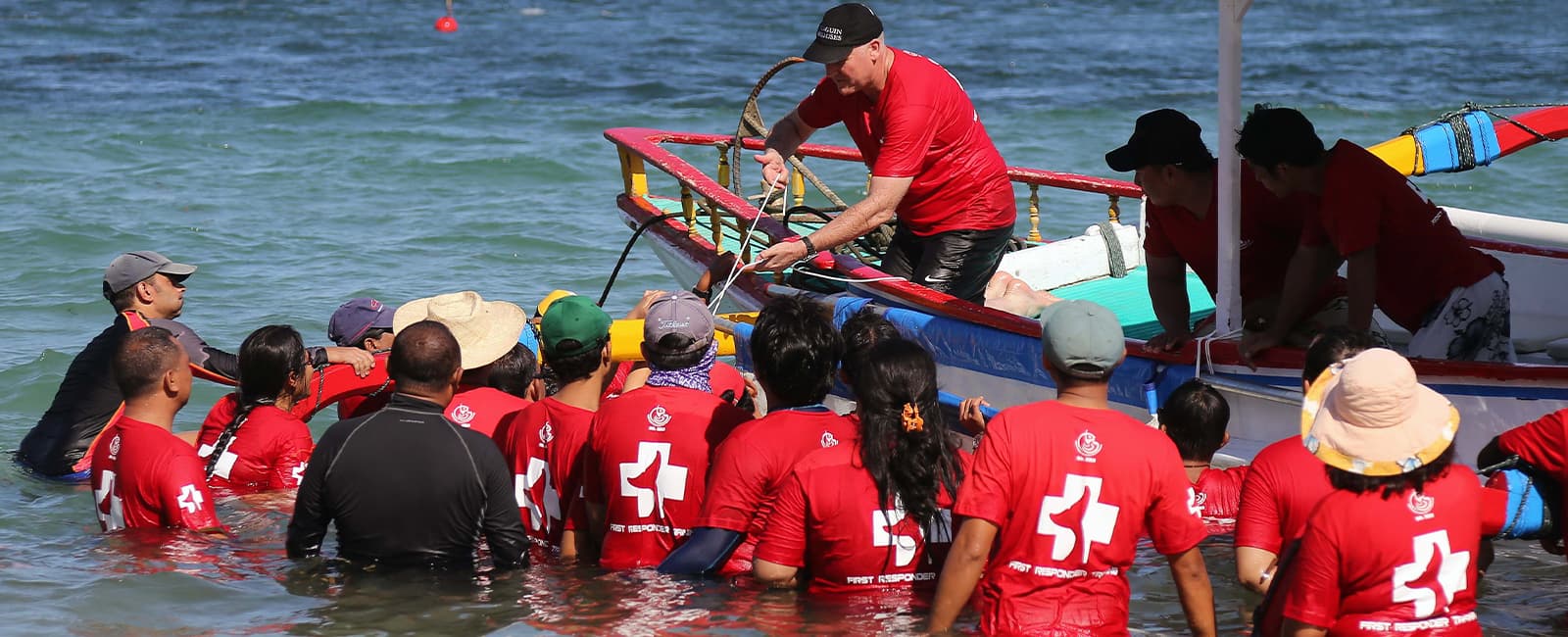 Many people in red shirts standing waist-deep in water around a boat.