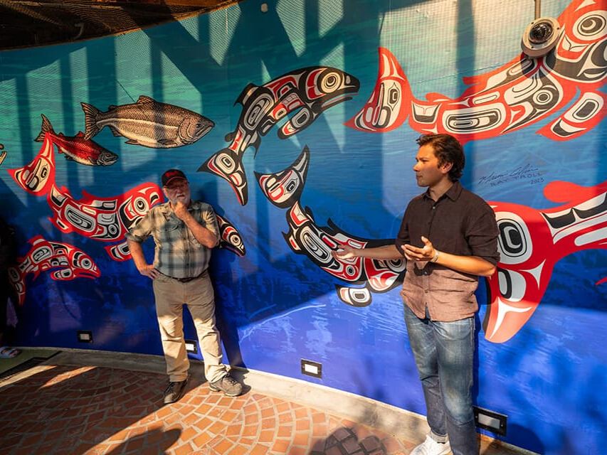 Ray Troll and Owen Oliver standing in front of a large blue mural depicting salmon.