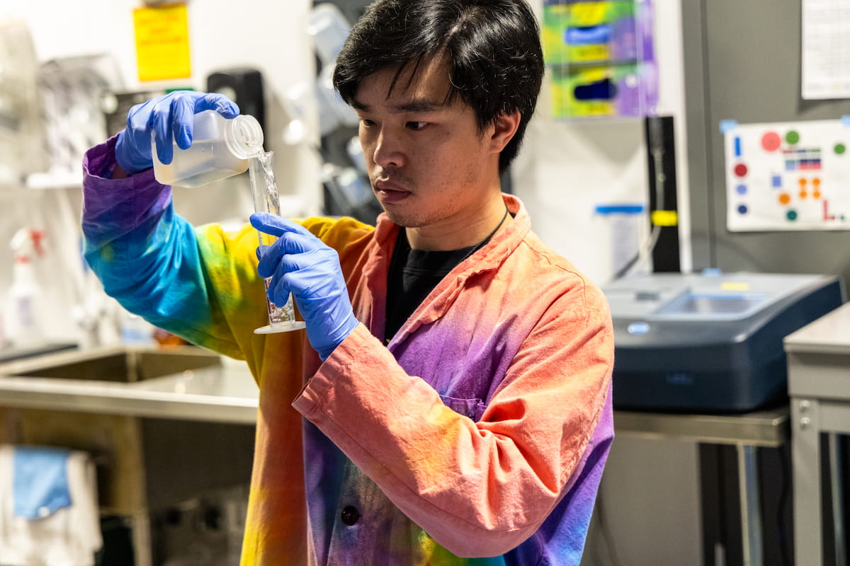 Keenan Wong pouring a solution into a graduated cylinder. Keenan wears a multicolored, tie-dyed lab coat.