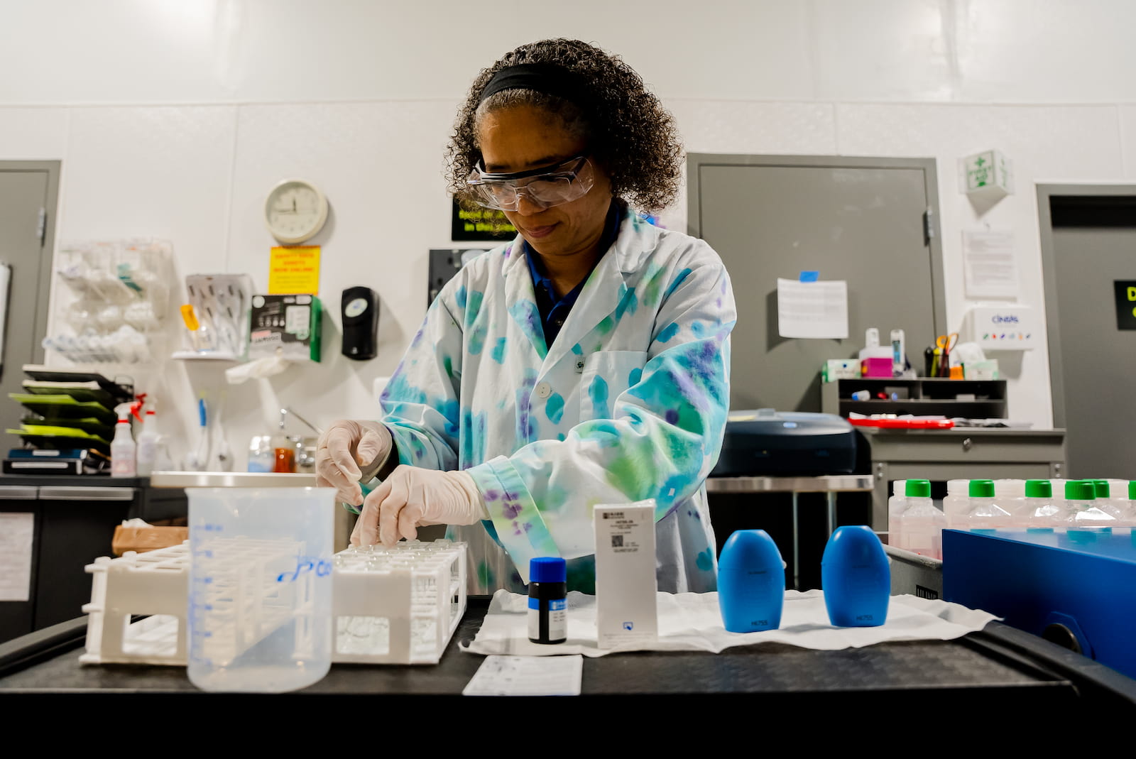 Angela Smith testing water quality samples in a lab. She wears safety glasses and a white lab coat speckled with blue and green.