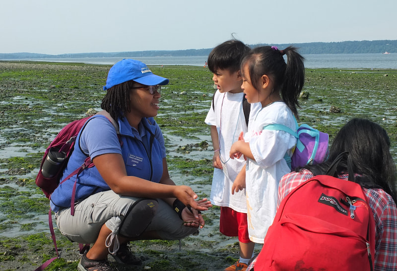 A Seattle Aquarium volunteer wearing a blue hat and shirt crouching on a beach. She is speaking to two children.